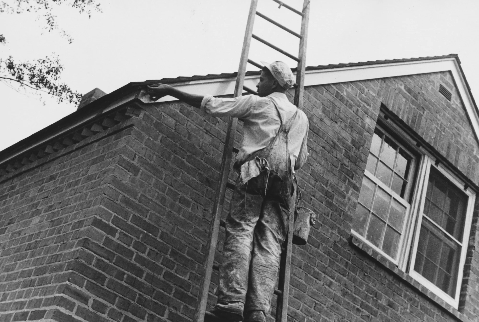 Black and white photo of a worker on a ladder at the eave of a brick house